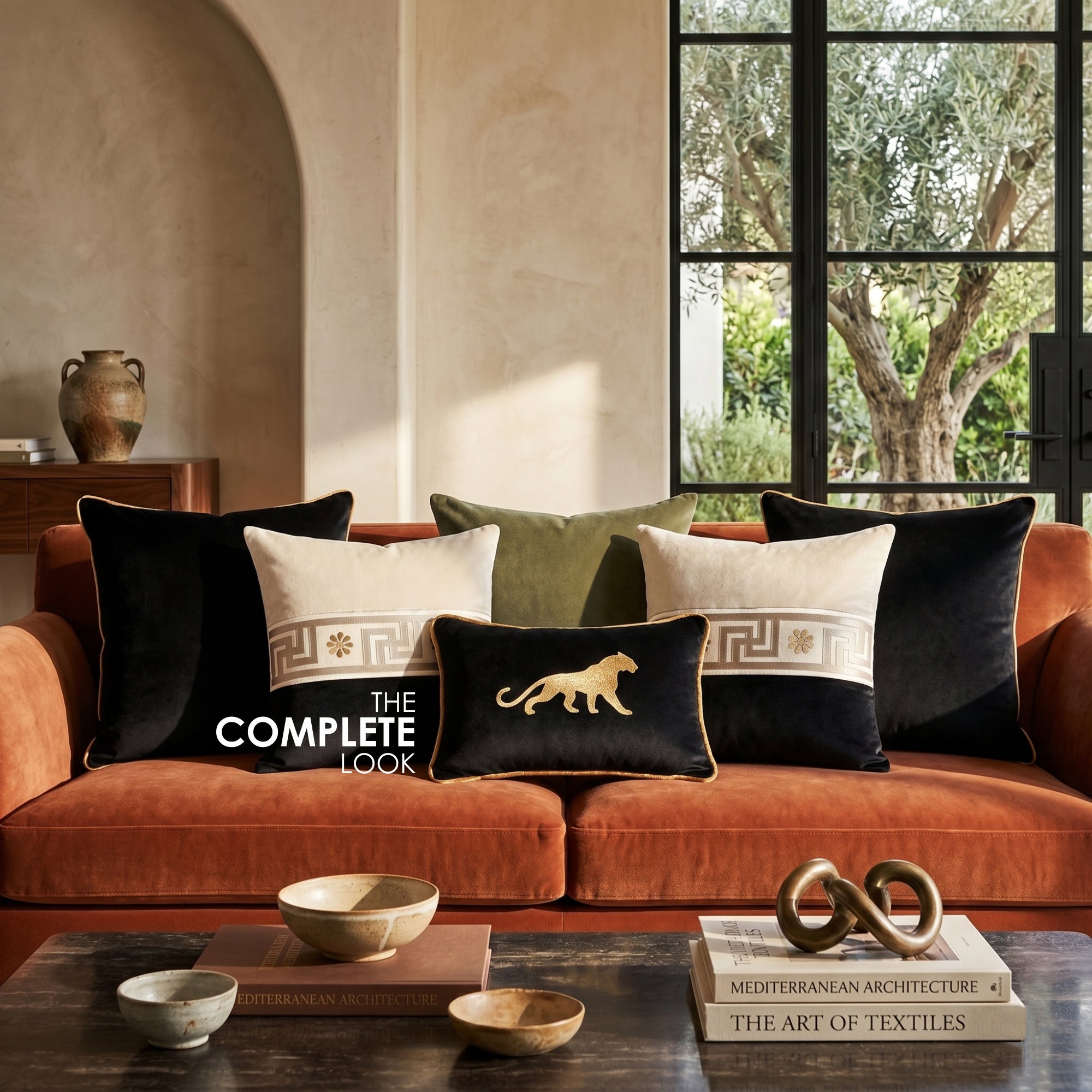 A styled living room scene showing an orange-brown velvet sofa with multiple decorative throw pillows. The pillows include black velvet with gold lion embroidery, cream and black Greek key patterned pillows, and an olive green pillow.